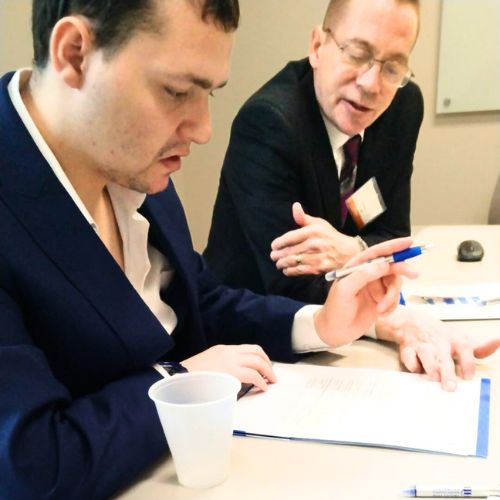 an older man in a suit helps a younger man in a suit jacket to fill out paperwork